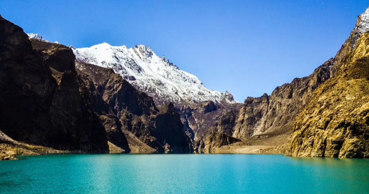 The vivid turquoise water of Attabad Lake below a snow-capped Karakoram peak and steep golden cliffs in Gojal, Upper Hunza, Gilgit-Baltistan, Pakistan