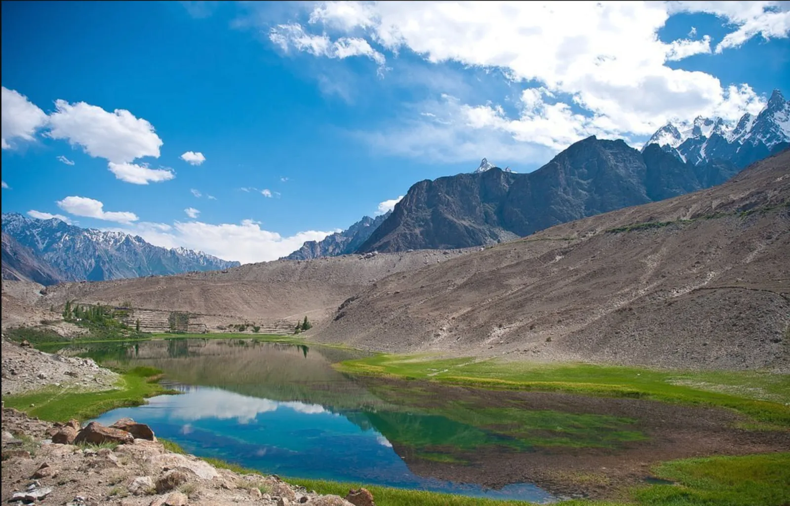 The still turquoise water of Borith Lake ringed by green meadow and barren Gojal slopes, Upper Hunza