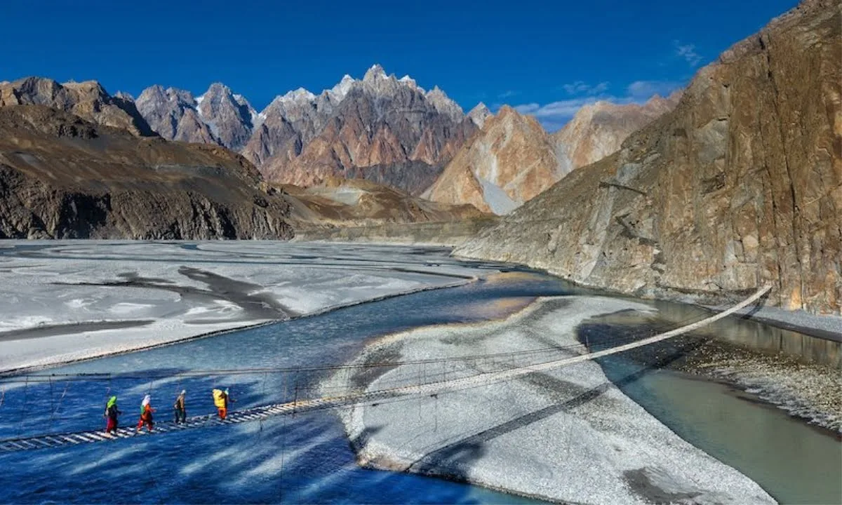 Trekkers crossing the slatted Hussaini Suspension Bridge over the Hunza River near Passu, with the jagged Passu Cones behind
